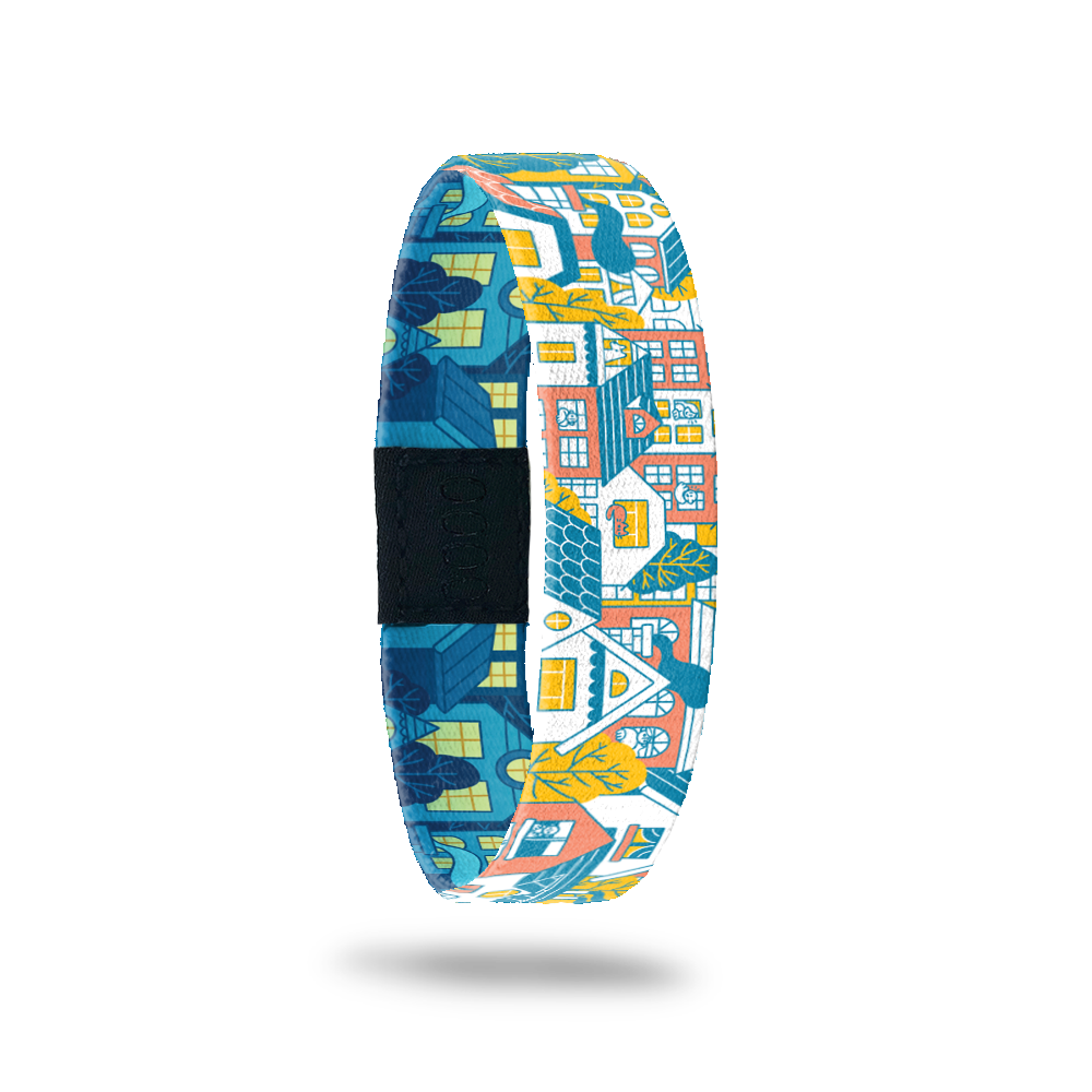 Wristband single with a design of a city block of houses and/or neighborhood. The colors are gold, light orange, blue and white. The inside is the same design but looks like nighttime so all of the houses are blue colored with glowing windows.The inside says Housing Is A Human Right. It comes with a matching charm. 