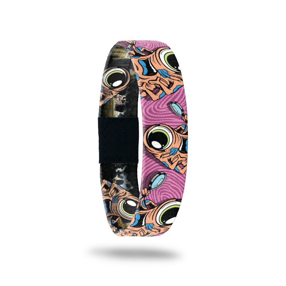 Pink swirly background with the monster in orange.  Monster is on the ZOX multiple times, has 1 eye, a blue tongue and a magnifying glass. Comes with a matching monster pin.