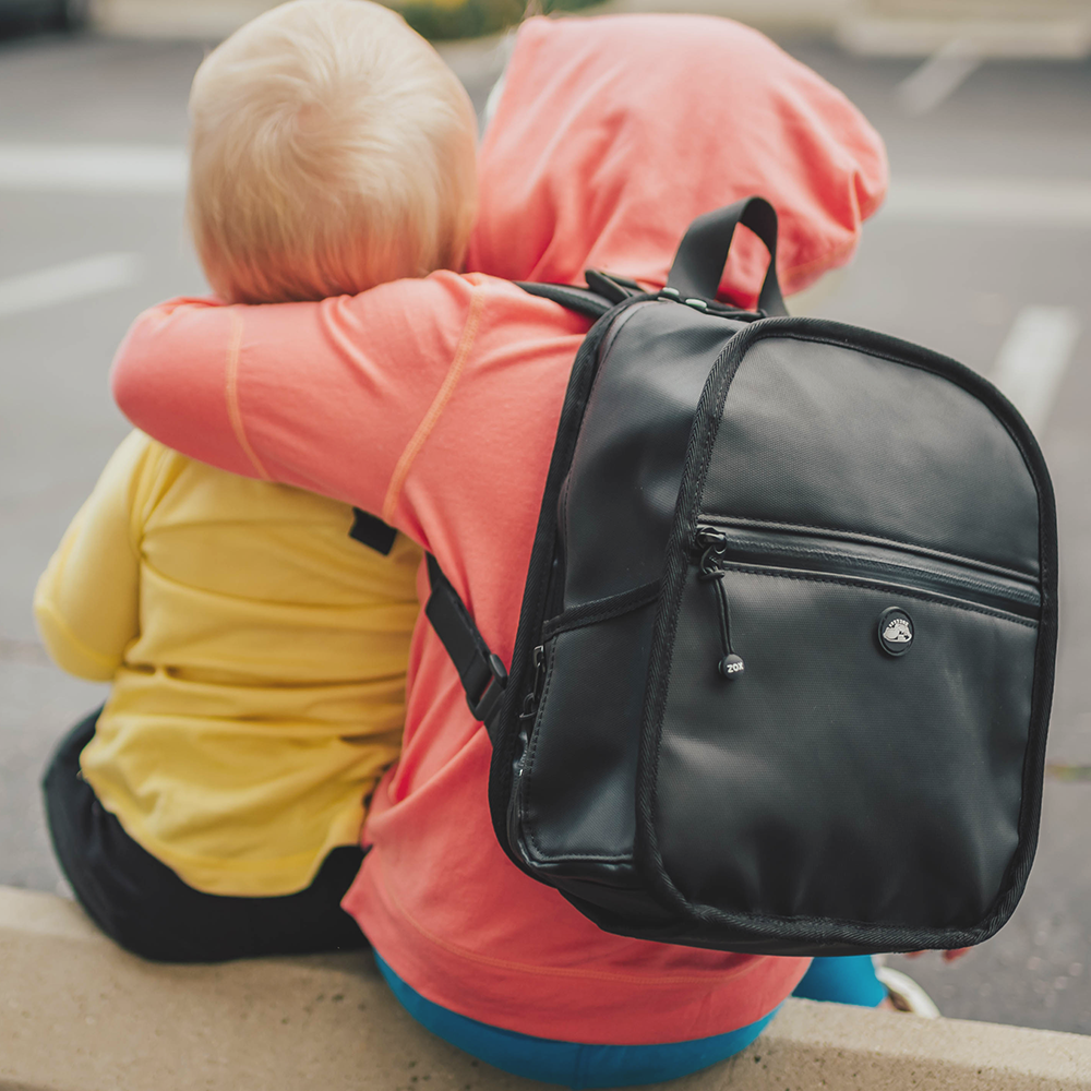 Lifestyle photo of a two little kids. Once is wearing the small black backpack while hugging the smaller little brother in the image