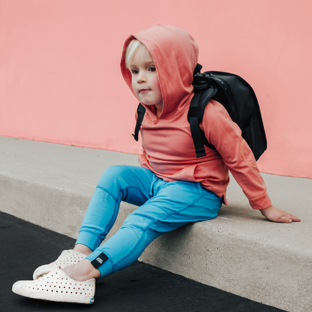 A lifestyle photo of a little boy wearing a pink and blue outfit and sitting on the ground with the small black backpack on his back.