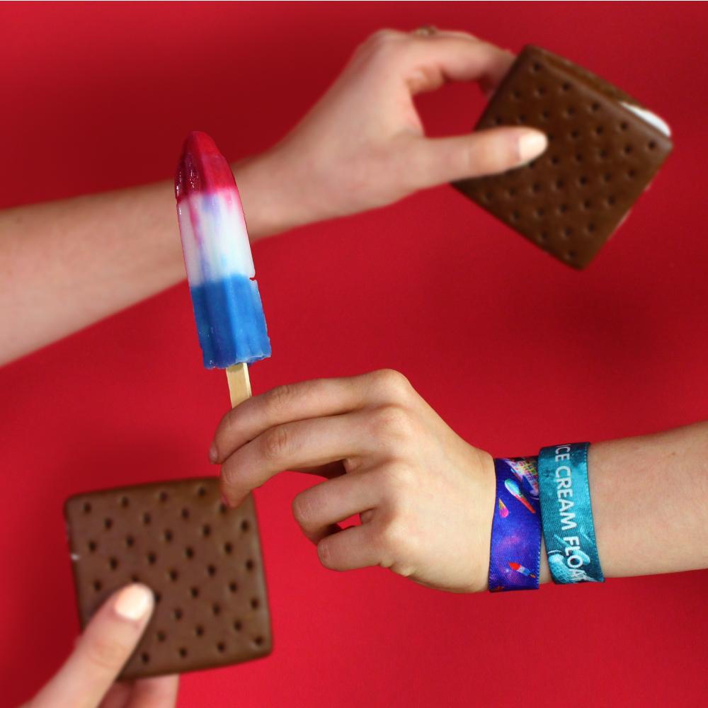 Studio photo of two Ice Cream Float straps on girls wrist holding a red white and blue popsicle and another girls hand holding some ice cream sandwiches