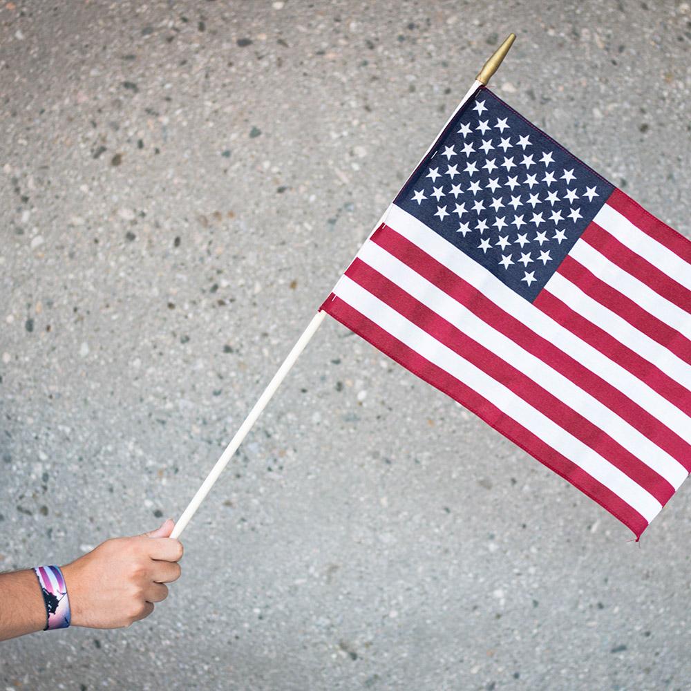 Lifestyle Image: hand holding United States flag with America, The Free wristband on their wrist