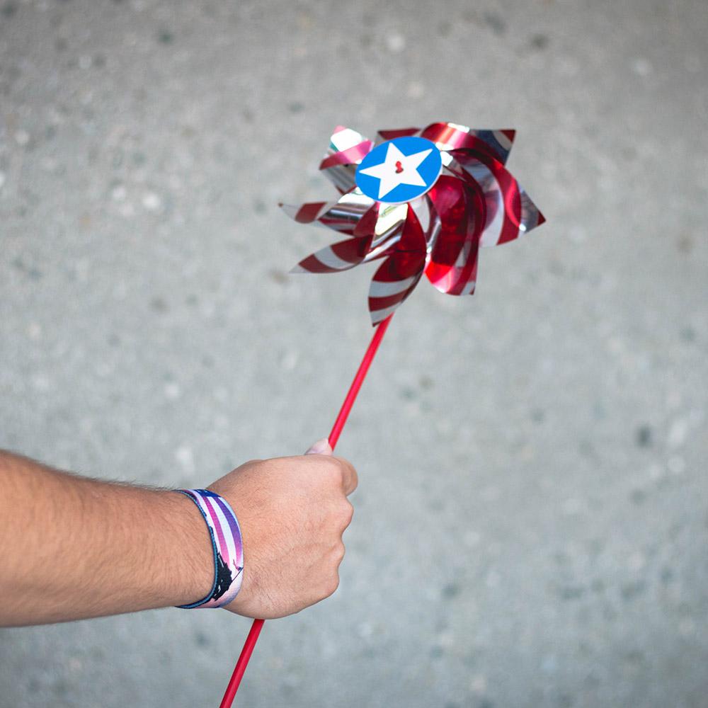 Lifestyle image of hand holding a red, white, and blue windmill with America, The Free on their wrist