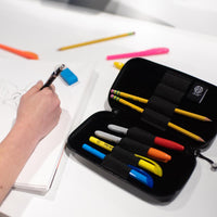 A lifestyle image showing the artist capsule open with markers and pencils in the spaces with a hand drawing on a notepad
