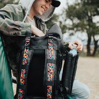 a man sitting down next to the imperial v2 backpack with the front showing so you are able to see the different tension straps that he has put in the bag, he is grabbing his water bottle from the water capsule that is attached to the bag.