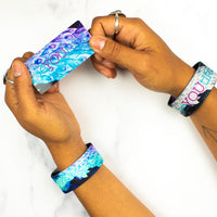 Studio image of hands with rings holding a card that says BeYOUtiful with one BeYOUtiful wristband on each wrist 