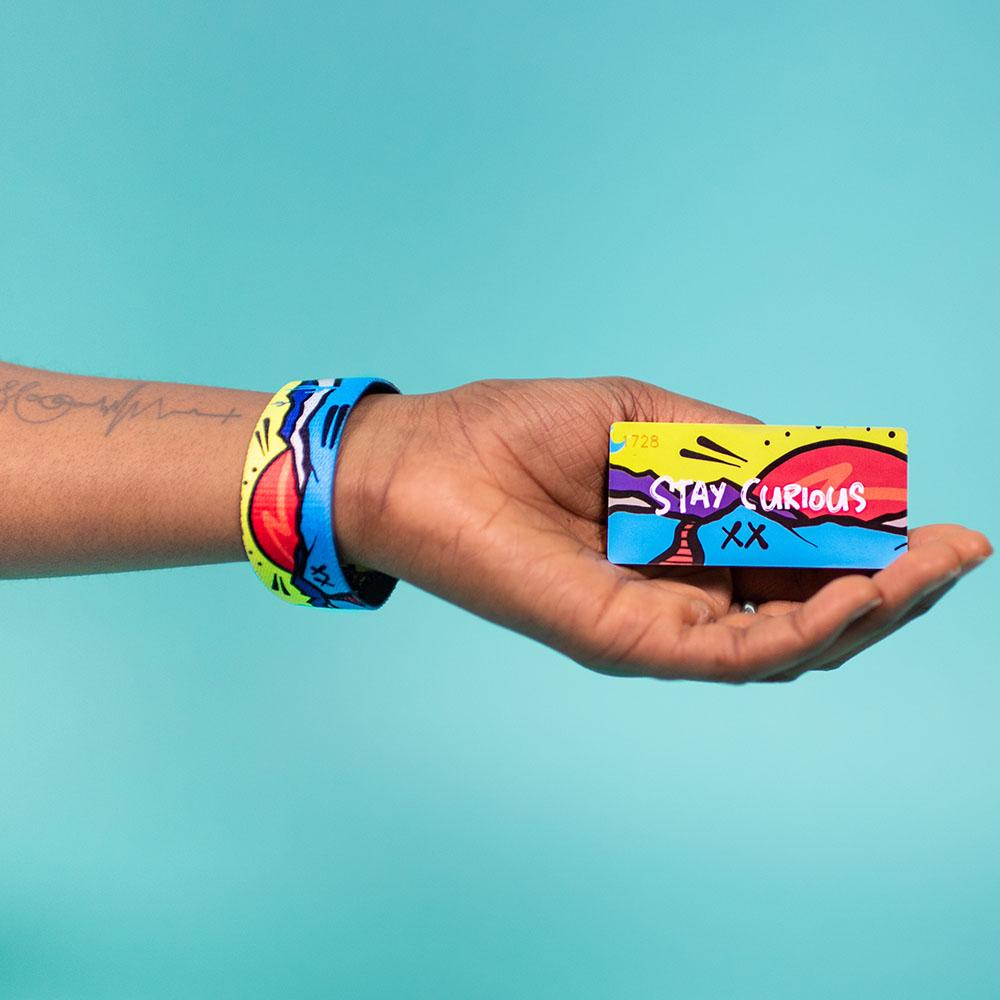 Studio Image of hand holding card that says Stay Curious with a Stay Curious strap on their wrist