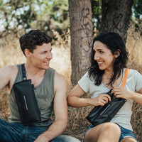 Life style image of a man and woman sitting down, the woman is unzipping the fanny pack to grab something from inside of it while the man has a water bottle holder around his shoulders.