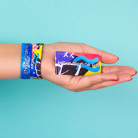 Studio photo of hand holding number-matching collector's card in front of bright blue background showing two fortune favors the bold singles one showing the outside design with hand drawn abstract art above another single with the inside design showing a bold white text fortune favors the bold with hand drawn abstract art