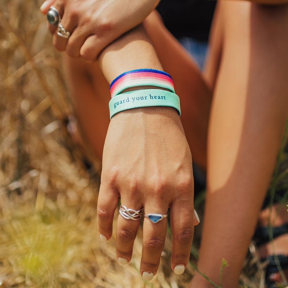 Lifestyle photo up close of girl's wrist wearing two guard your heart singles showing the outside design of guard your heart with mint, green, peach, pink, red, maroon, purple, blue rainbow design above another showing the  inside design with black text guard your heart with varying background in mint, green, peach, pink, red, maroon, purple, or blue