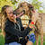 girl smiling in black hoodie and 2 heart of gold wristbands petting a grayhound. 