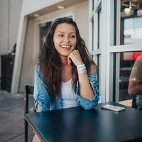 Lifestyle image of woman sitting at a table smiling and wearing 2 Listen wristbands