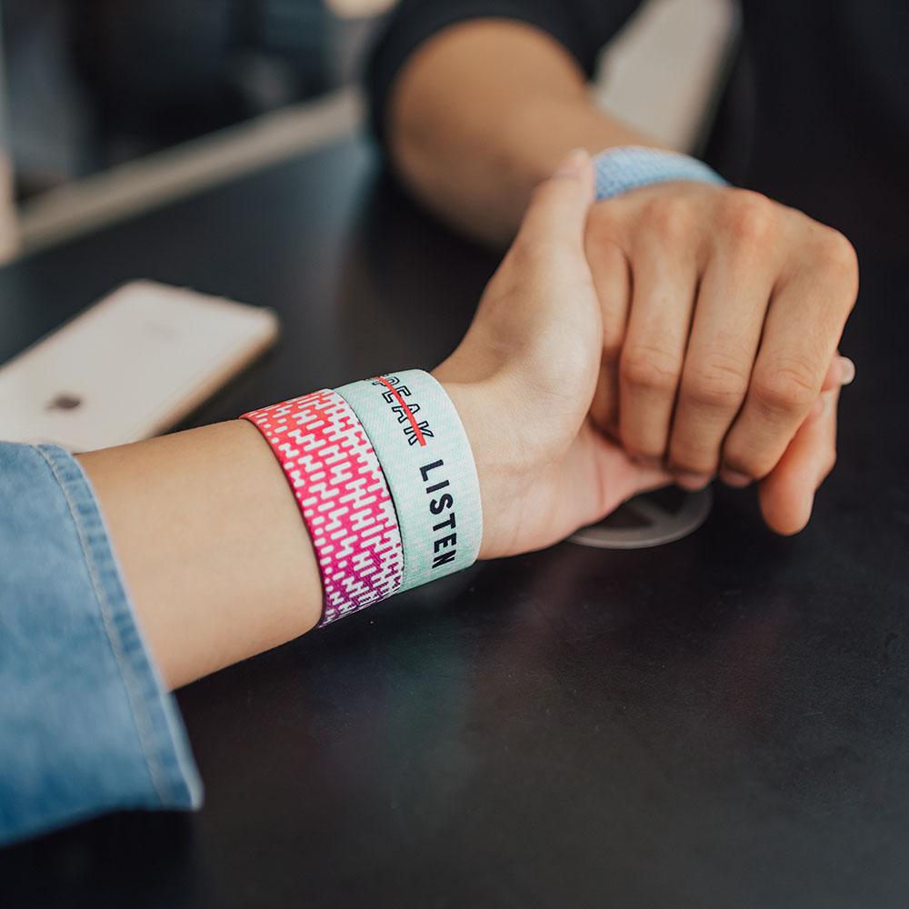 Lifestyle image of hands holding on a table. One has 2 Listen wristbands and the other has 1 Listen wristband on