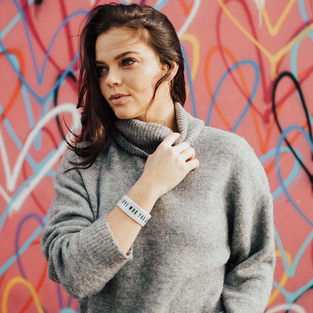 Lifestyle image of a woman in front of a colorful background wearing a grey sweatshirt and Love Who You Are on her wrist