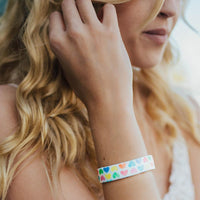 Lifestyle photo up close of girl showing wrist with live with love single showing the outside design of with rows of pastel orange, yellow, pink, green, purple, and blue gradient hearts over white background