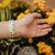 Lifestyle photo of hand reaching for flowers showing wrist with live with love single showing the outside design of with rows of pastel orange, yellow, pink, green, purple, and blue gradient hearts over white background