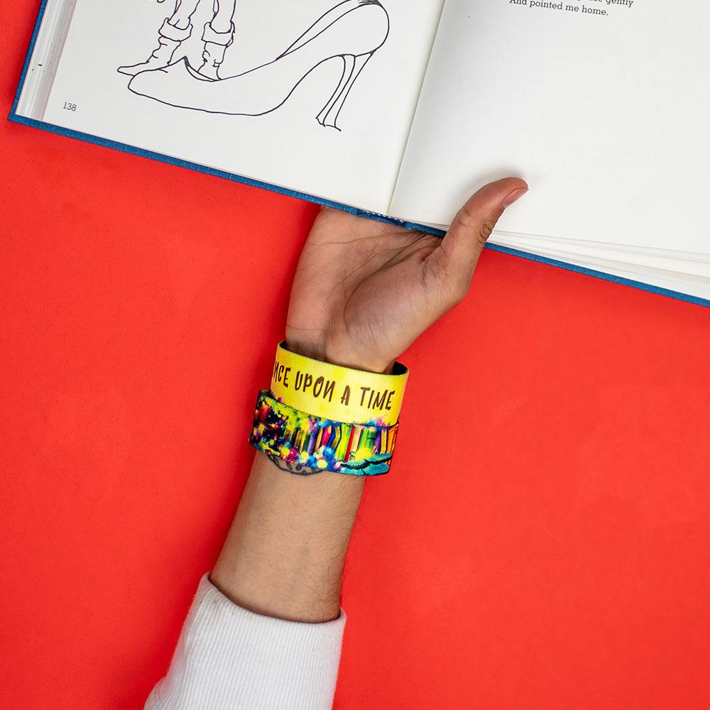 Studio image of hand holding an open book and wearing 2 Once Upon A Time on their wrist. 