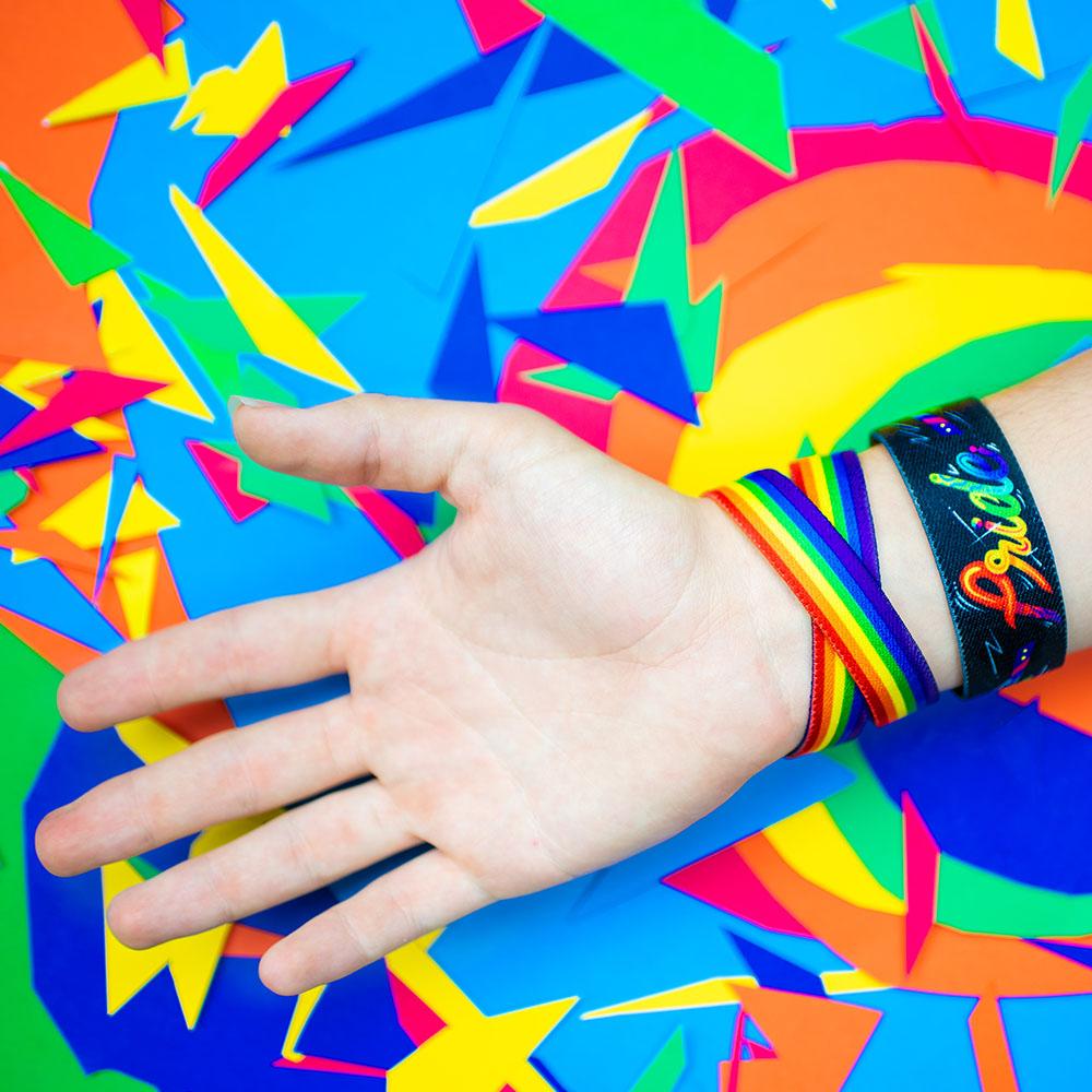 Studio image close up of Pride and a double wristband called Love Wins on someone's wrist in front of a colorful background