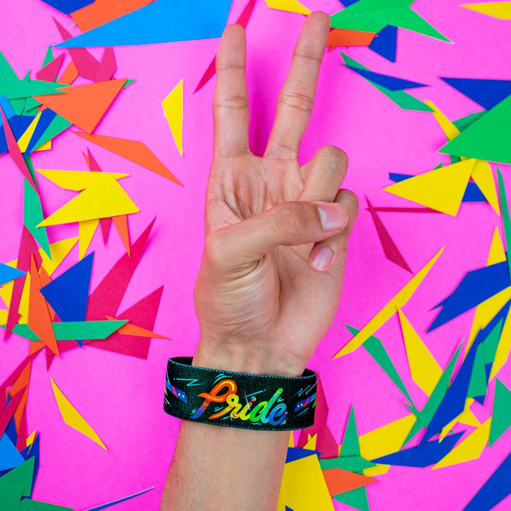Studio close up image of a hand giving the peace hand sign in front of a colorful background and wearing a Pride strap