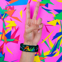 Studio close up image of a hand giving the peace hand sign in front of a colorful background and wearing a Pride strap