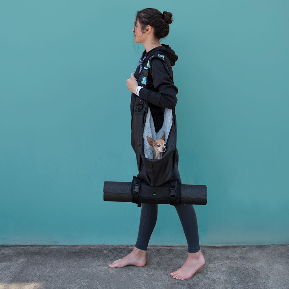A woman walking with a dog carrier with the roll connected to the bottom that is holding a yoga matt