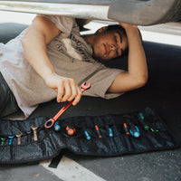 lifestyle image of a man under his car fixing it with the roll open near him full of tools in the pockets
