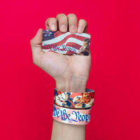 Studio image of hand holding a card that says We The People and wearing 2 We The People on their wrist